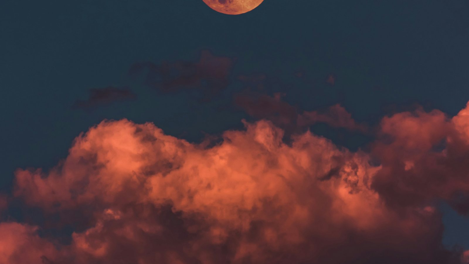 Lune orangée à demi sortie du cadre au-dessus d'un banc de nuages teintés d'orange, dans un ciel bleu sombre de fin de nuit
