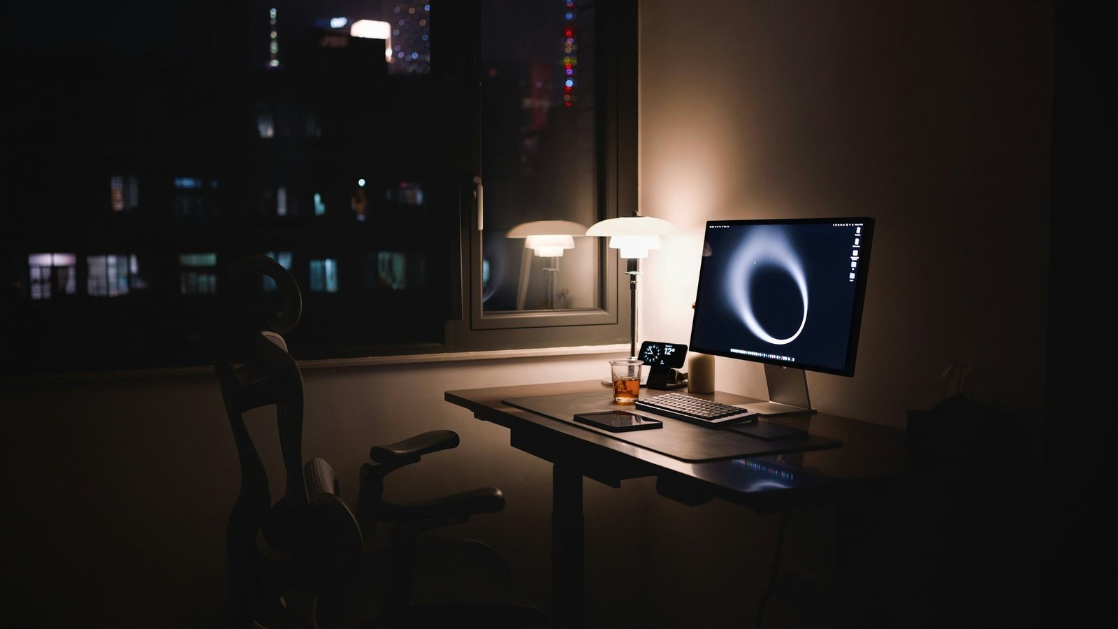Bureau vide éclairé par une lampe le soir, écran allumé, fenêtre sur la ville