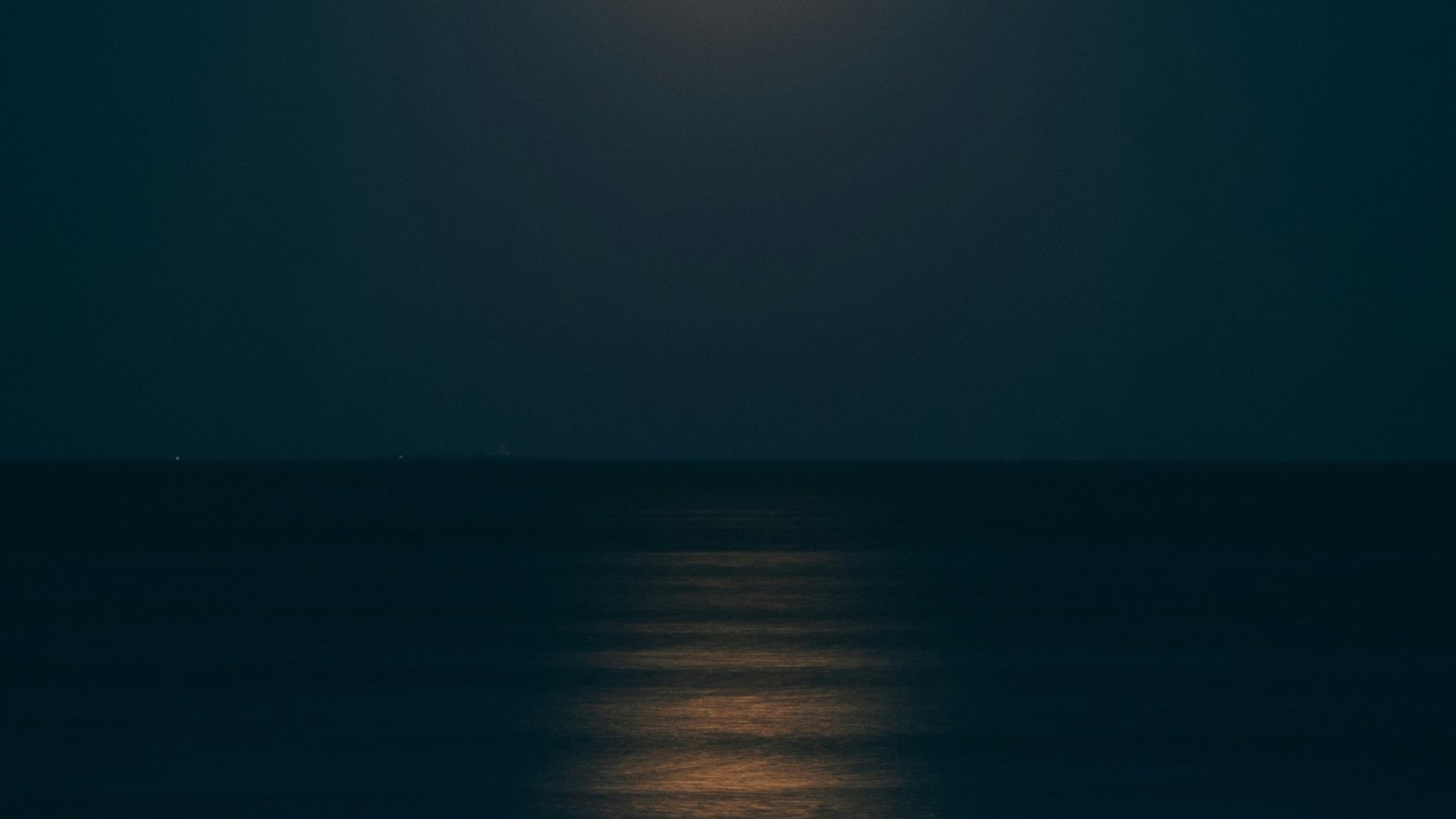 Mer sombre la nuit sous un ciel bleu nuit, avec le reflet doré de la Lune tracé sur l'eau et une ligne d'horizon entièrement dégagée