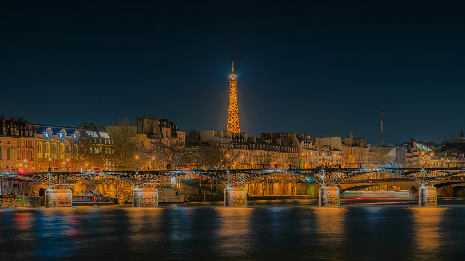 Berges d'un fleuve dans une grande ville la nuit, avec un pont métallique éclairé au premier plan, des immeubles alignés et une tour illuminée qui domine l'horizon