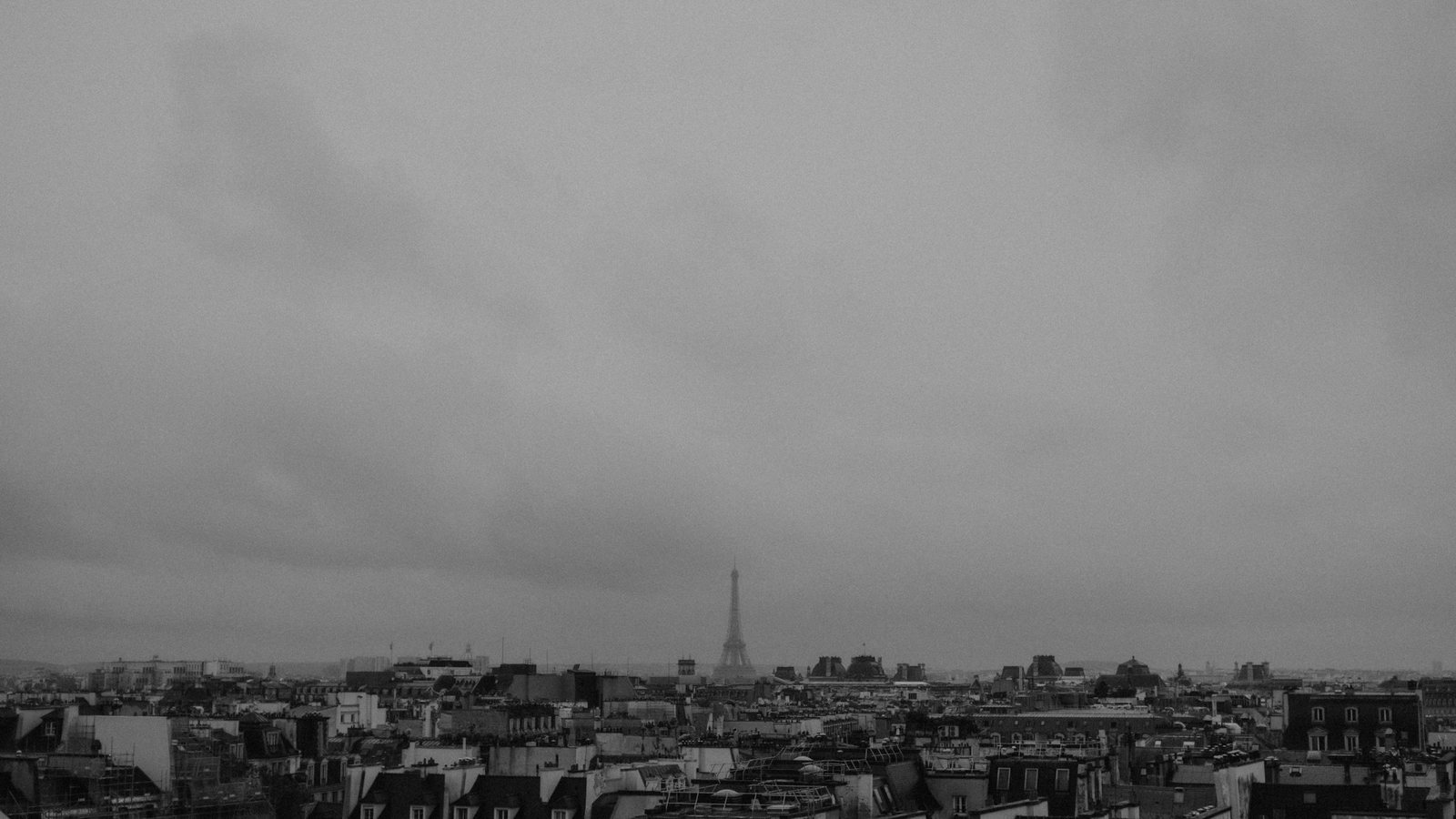 Toits d'immeubles urbains en noir et blanc sous un ciel entièrement couvert, la tour Eiffel se devinant au loin sur l'horizon