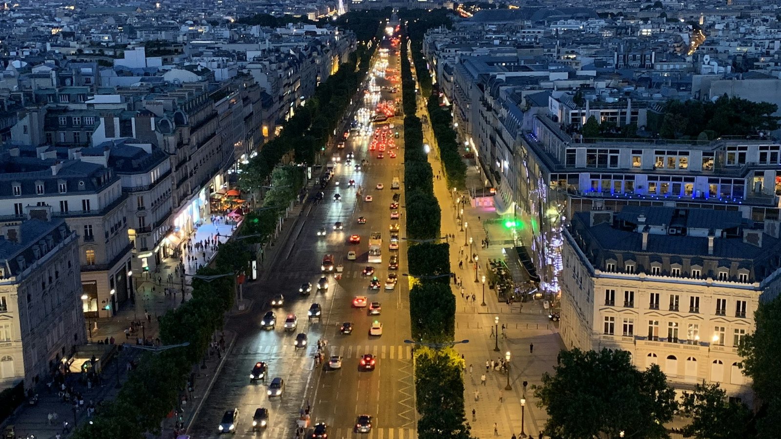 Vue plongeante sur une large avenue urbaine à la tombée de la nuit, bordée d'immeubles alignés et de rangées d'arbres, avec des files de voitures aux phares allumés et l'éclairage public en fonctionnement