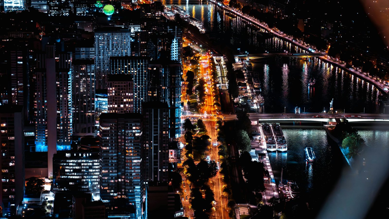 Vue aérienne nocturne d'une grande ville traversée par un fleuve, avec des quais éclairés, des ponts, des péniches amarrées et des immeubles de grande hauteur allumés
