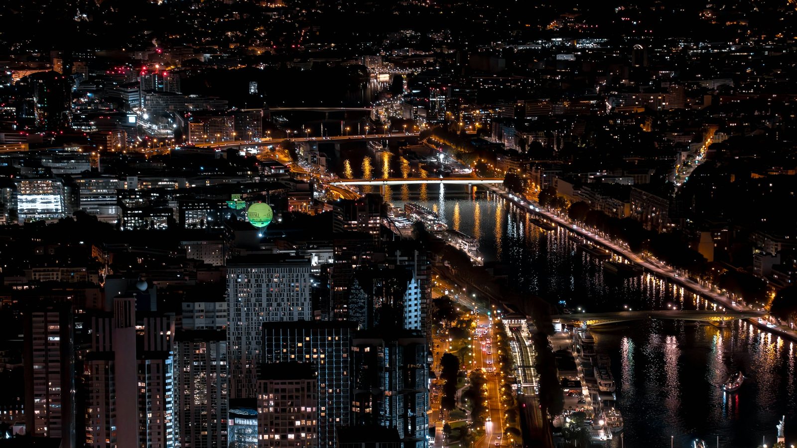 Vue aérienne d'une grande ville la nuit, avec des quais éclairés le long d'un cours d'eau franchi par plusieurs ponts, des tours d'habitation et des boulevards illuminés