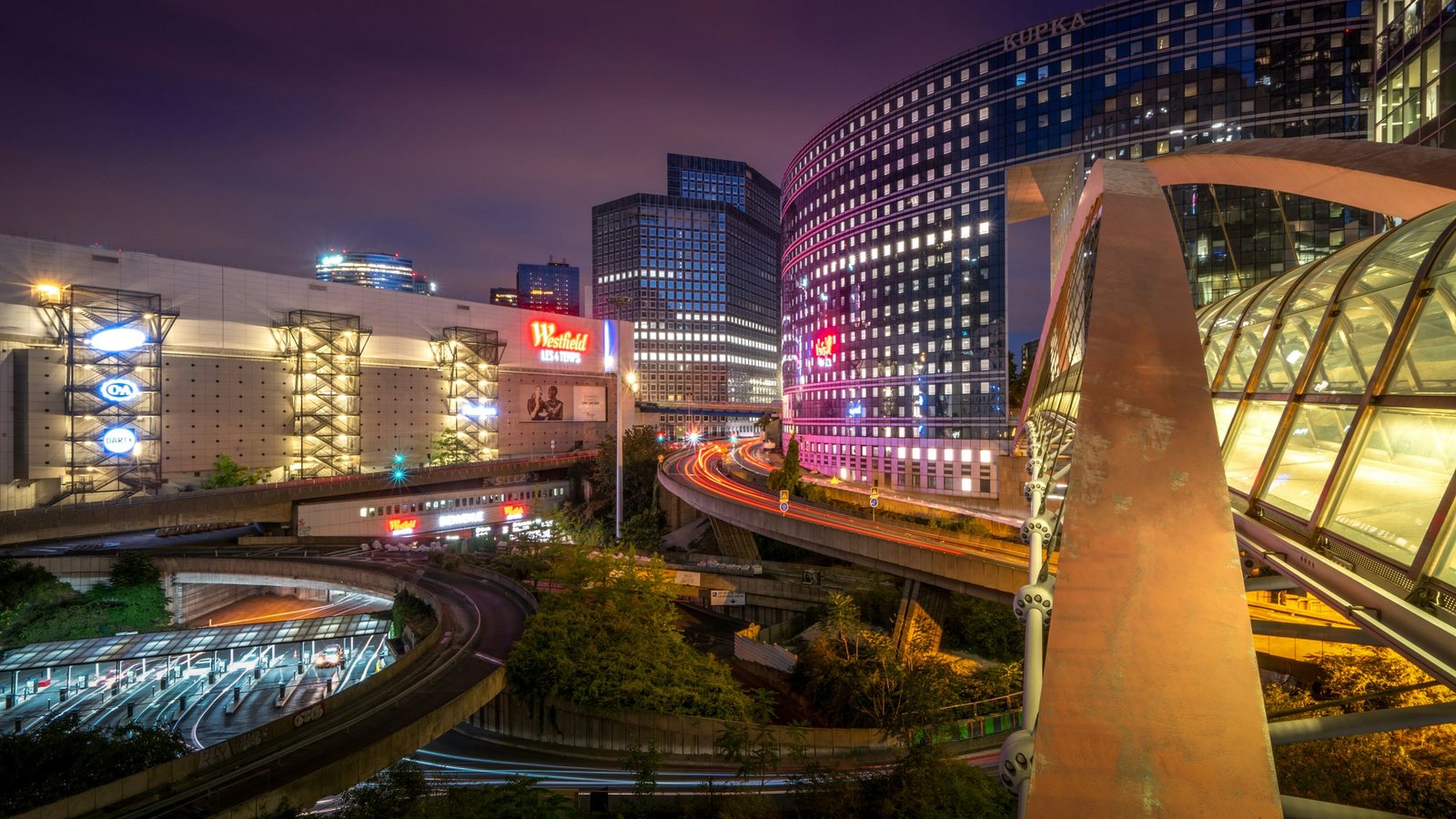 Quartier urbain photographié de nuit : tours de bureaux éclairées, façades commerciales lumineuses, échangeur routier à plusieurs niveaux et traînées de phares, sans la moindre ouverture vers l'horizon