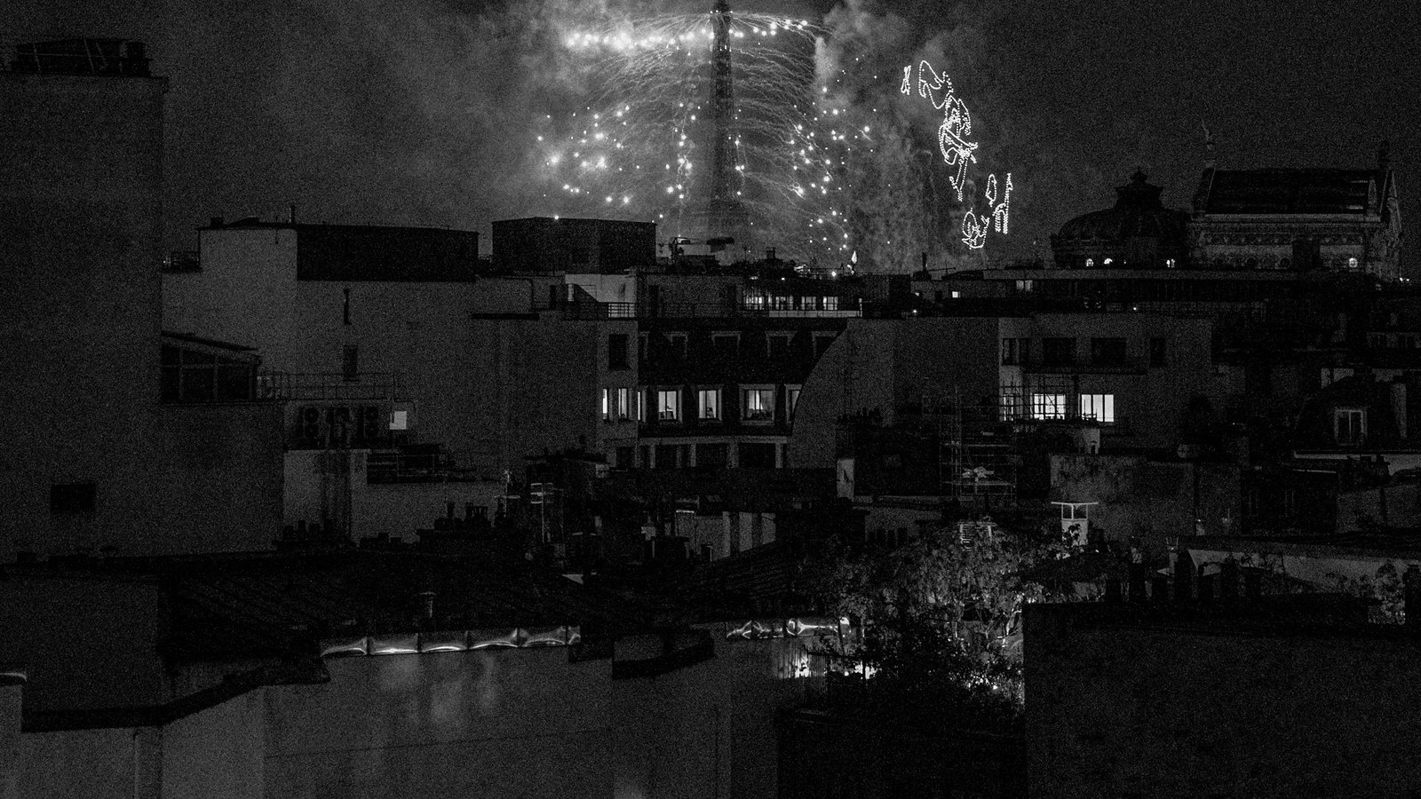 Toits d'une grande ville photographiés de nuit en noir et blanc, cheminées et immeubles serrés devant un ciel sombre où brille un feu d'artifice derrière une tour au loin