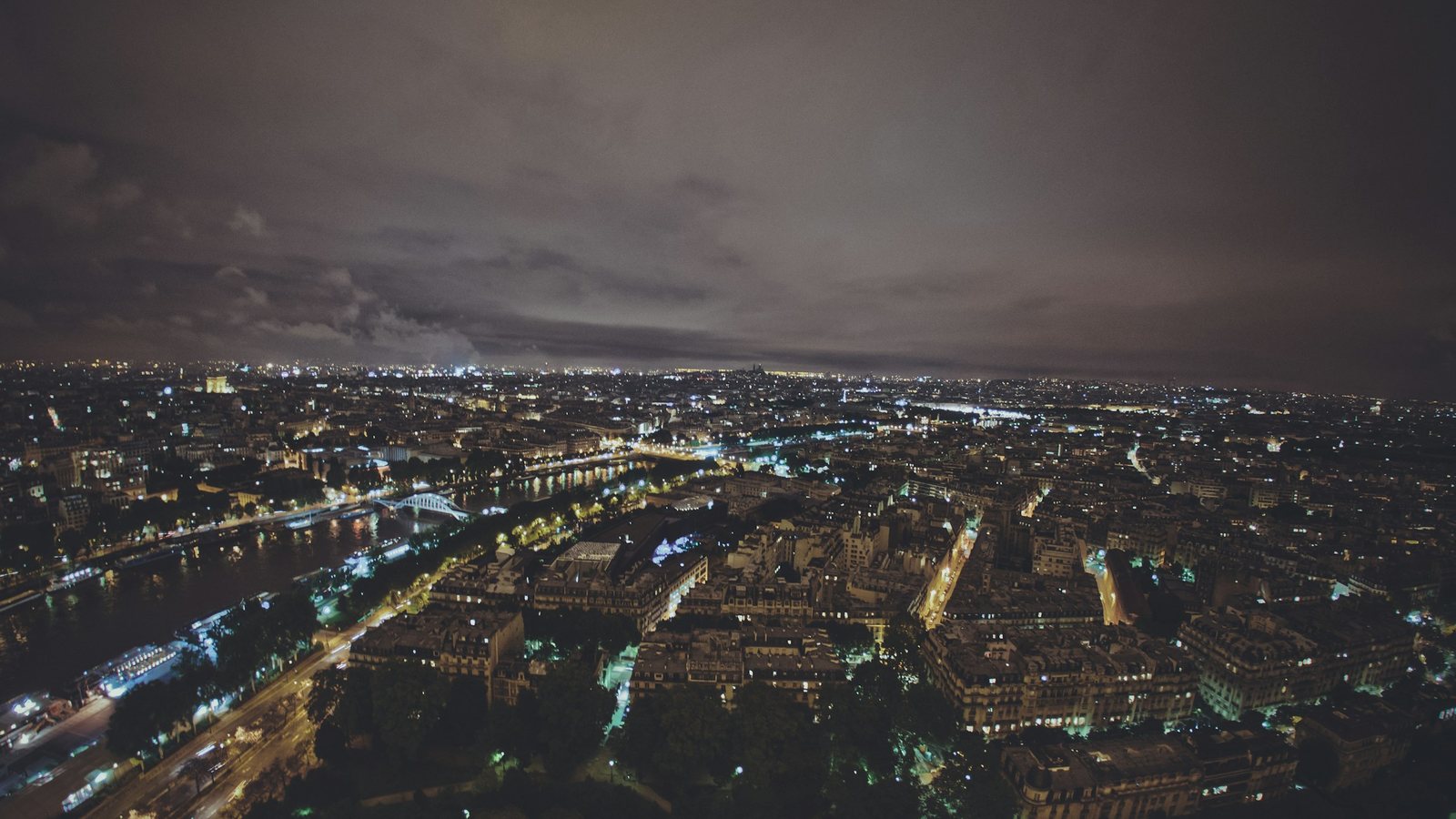 Vue aérienne nocturne d'une grande ville traversée par un fleuve et ses ponts, toits serrés et boulevards éclairés sous un plafond de nuages bas