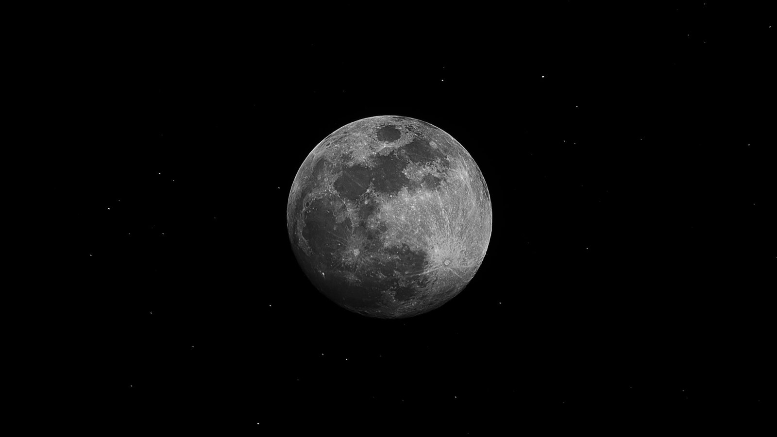 Disque lunaire presque plein photographié en noir et blanc, mers sombres et cratères bien visibles, sur un fond de ciel noir semé de quelques points lumineux