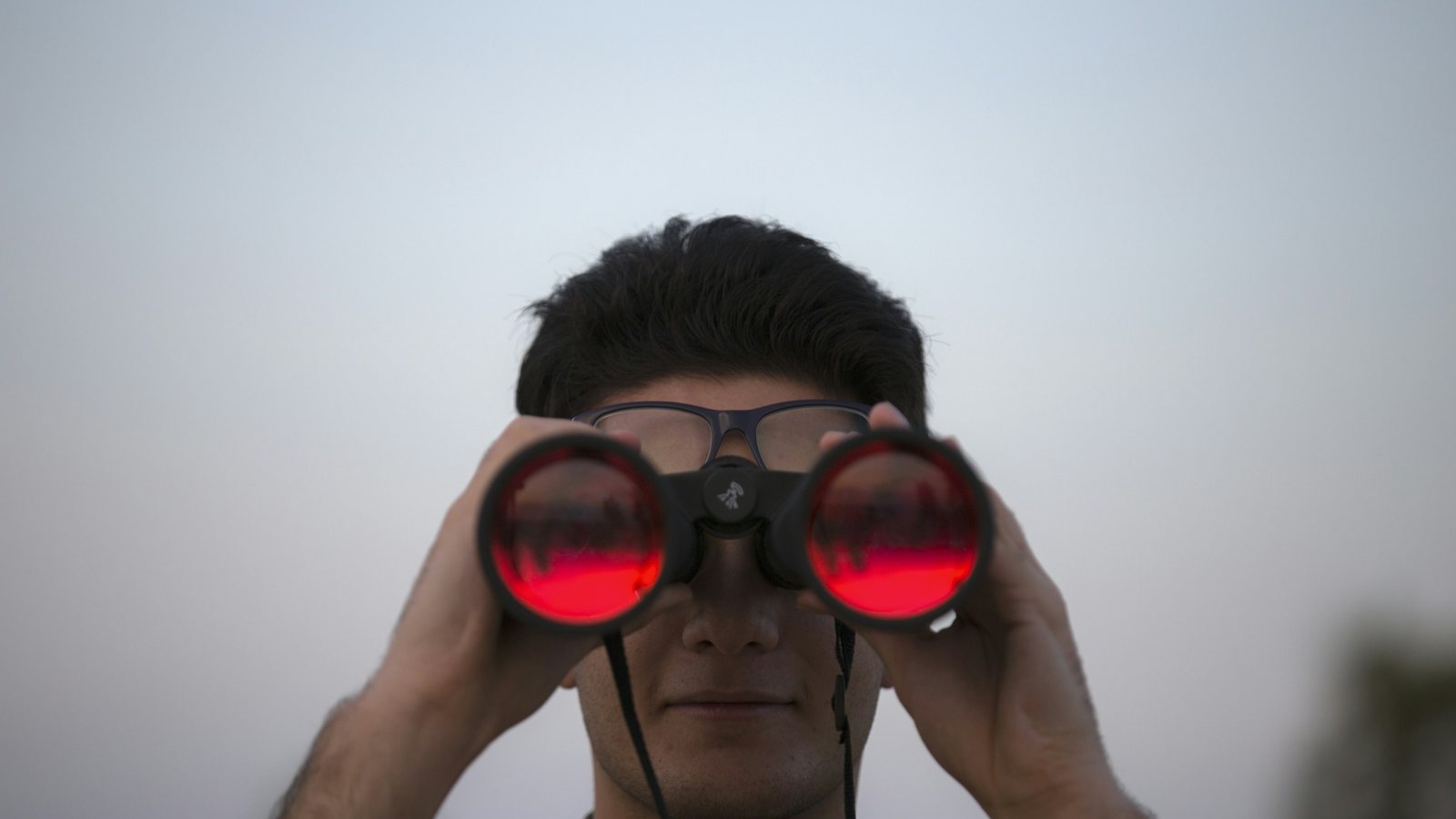 Homme de face tenant à deux mains une paire de jumelles noires devant ses yeux, les objectifs renvoyant un reflet rouge, sur un fond de ciel gris uni