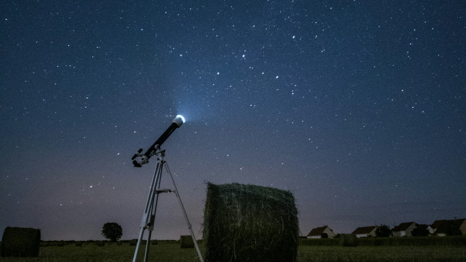 Petite lunette astronomique installée sur un trépied dans un champ moissonné, à côté d'une botte de foin ronde, sous un ciel étoilé avec quelques maisons au loin sur l'horizon