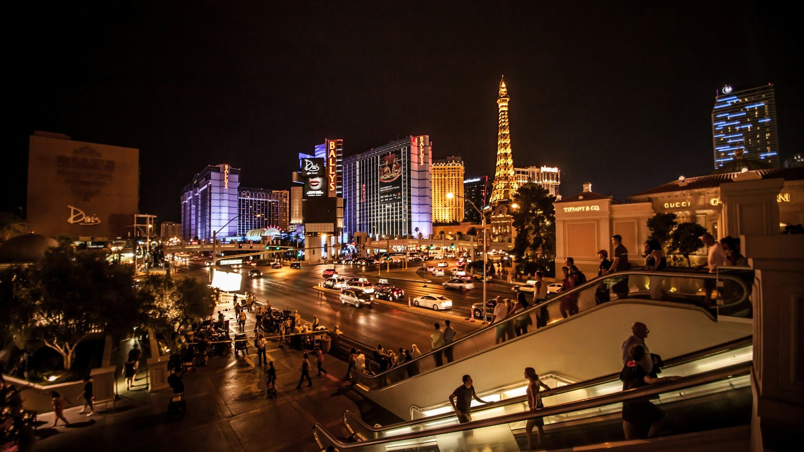 Grande avenue urbaine de nuit saturée d'éclairages artificiels, façades illuminées, passants et voitures, sans aucune portion de ciel dégagée au-dessus des bâtiments