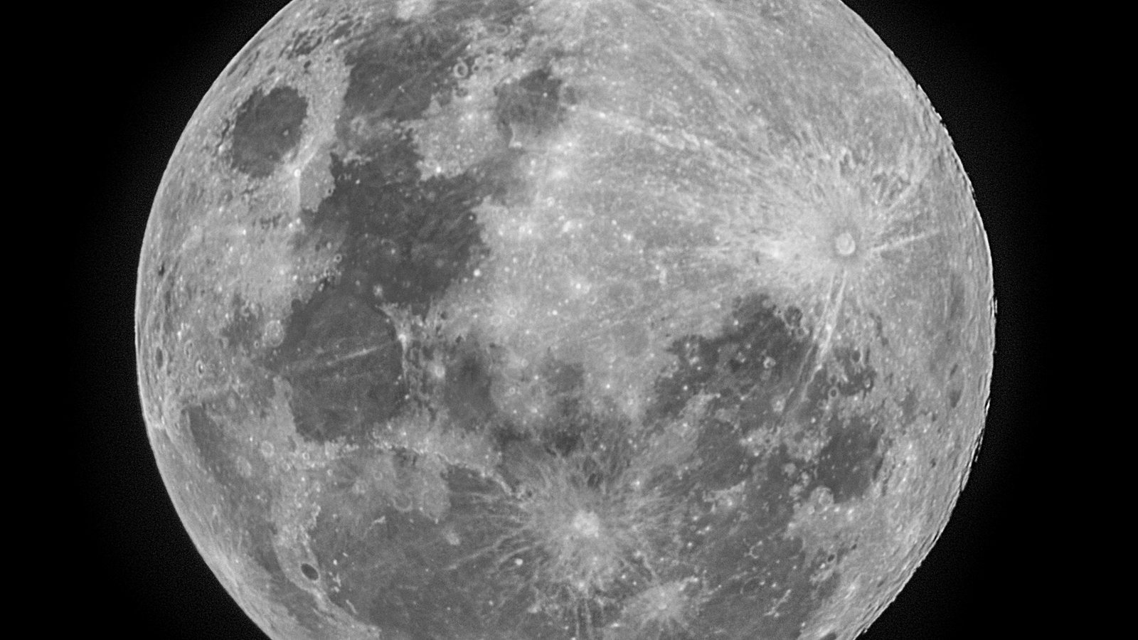 Gros plan en noir et blanc d'une Lune presque pleine sur fond de ciel noir, avec ses mers sombres, ses cratères et les longues traînées claires qui en rayonnent