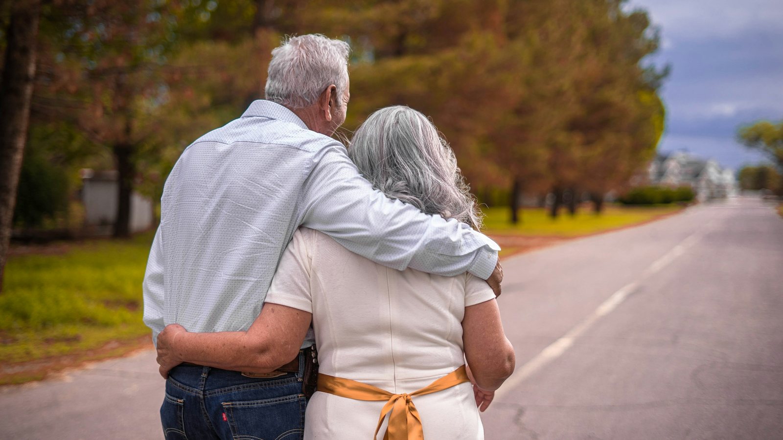 Couple âgé aux cheveux blancs marchant enlacé sur une route, photographié de dos