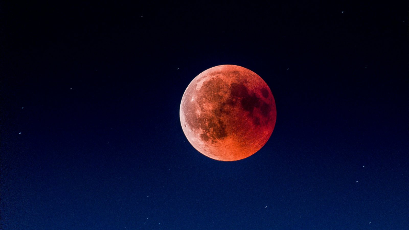 Disque lunaire entièrement plongé dans l'ombre, teinté de rouge cuivré, sur un ciel nocturne bleu très sombre parsemé de quelques étoiles